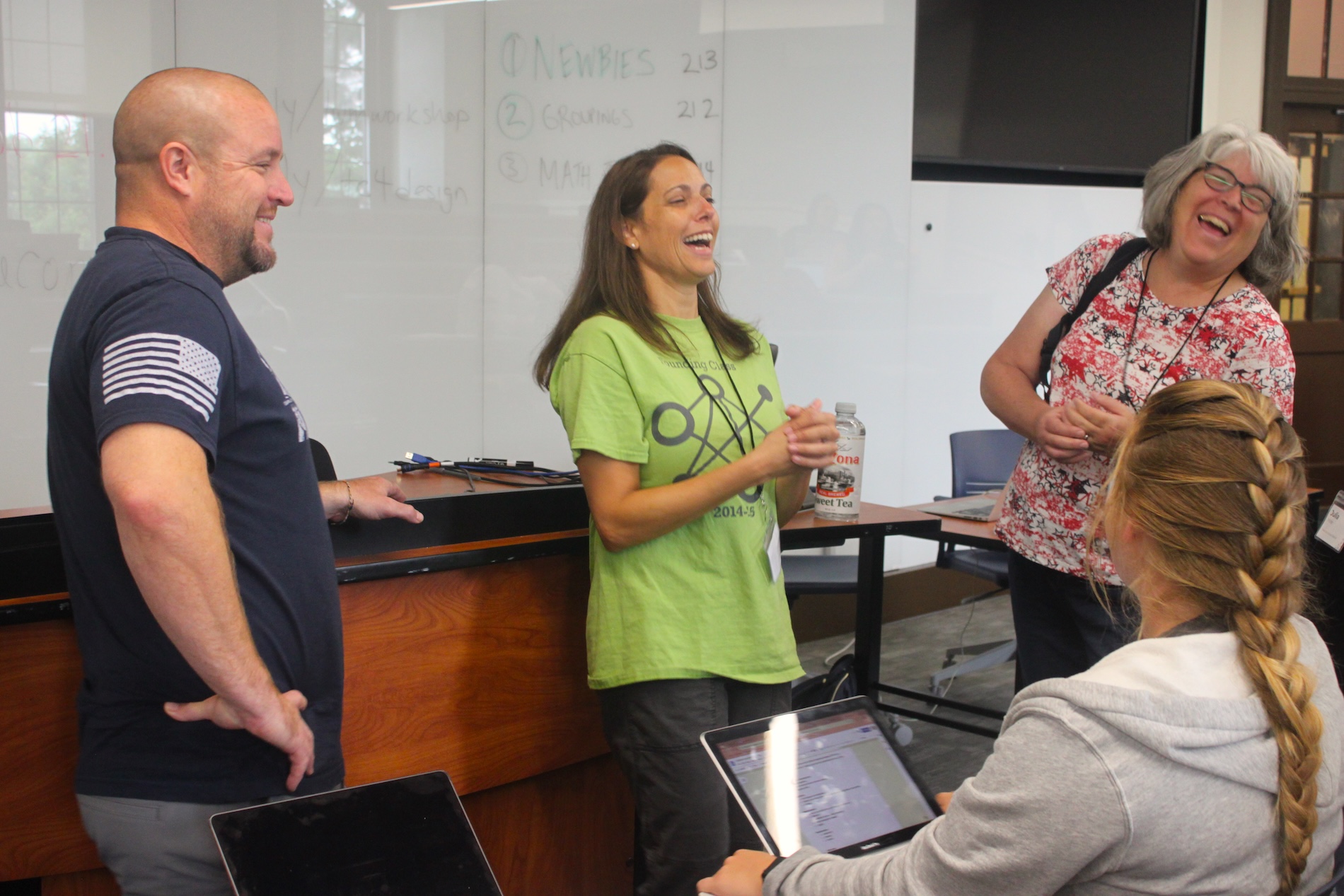 A group of four adults laugh and talk together in a classroom setting, gathered near a whiteboard and desks, with one person seated using a laptop.