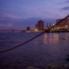 A twilight view of the New Orleans skyline with city lights reflecting on the Mississippi River, a bridge illuminated in the distance, and a chain in the foreground.