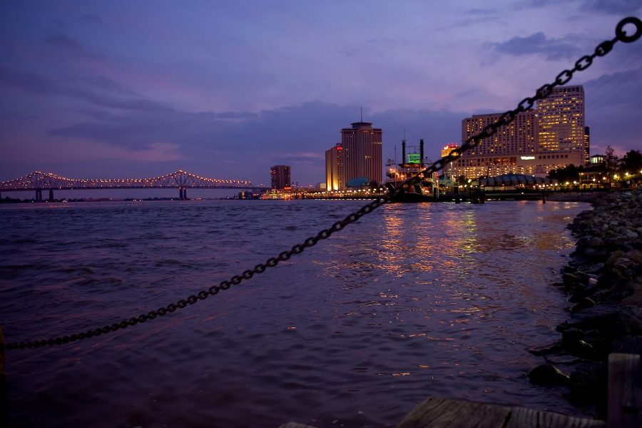 A twilight view of the New Orleans skyline with city lights reflecting on the Mississippi River, a bridge illuminated in the distance, and a chain in the foreground.
